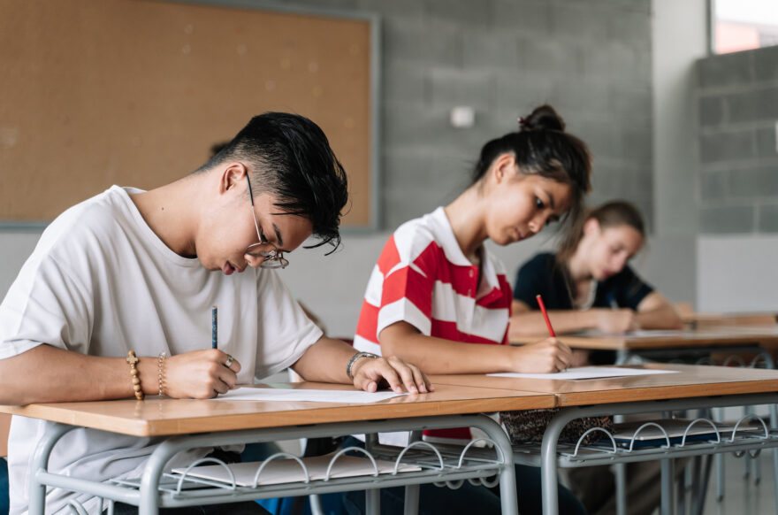 A student sits at a desk in a classroom, focused on writing in a notebook. Other students and a whiteboard with lesson notes are visible in the background.