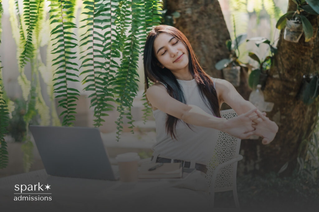A woman stretching her arms above her head while seated in front of a laptop on an outdoor desk setup, surrounded by greenery and natural light, symbolizing relaxation and work-life balance.