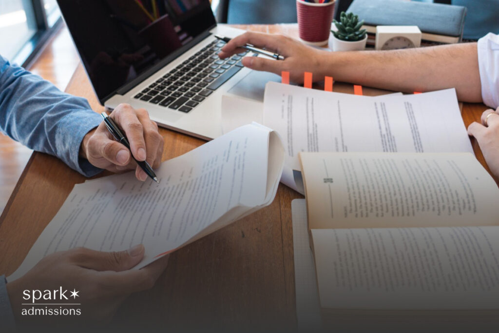 A table with documents spread out, featuring the hands of a professor and a student. The professor’s hand gestures toward a specific document, indicating guidance, while the student’s hand holds a pen poised to take notes. The table surface is wooden, and the documents include charts, handwritten notes, and printed materials.
