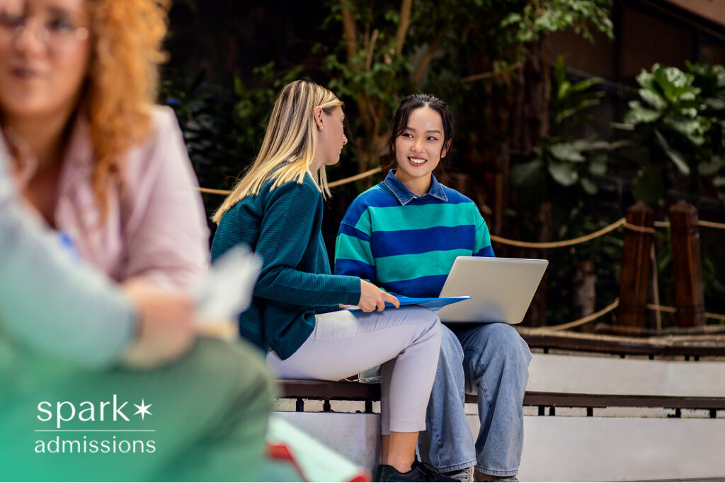 Two college students sitting outdoors on campus, smiling and working together on a laptop, representing collaborative learning and campus life.