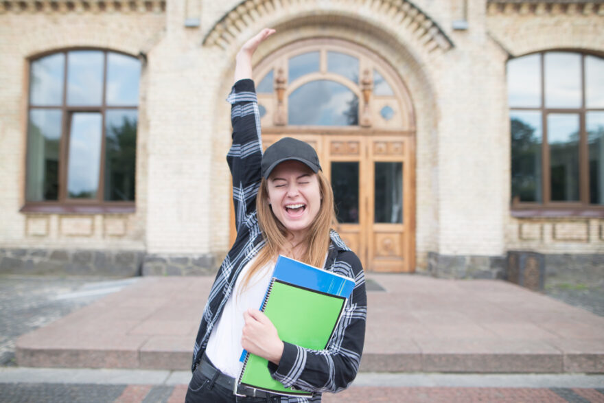 Pretty girl stands on the background of university with books and notebooks in her hands and rejoices